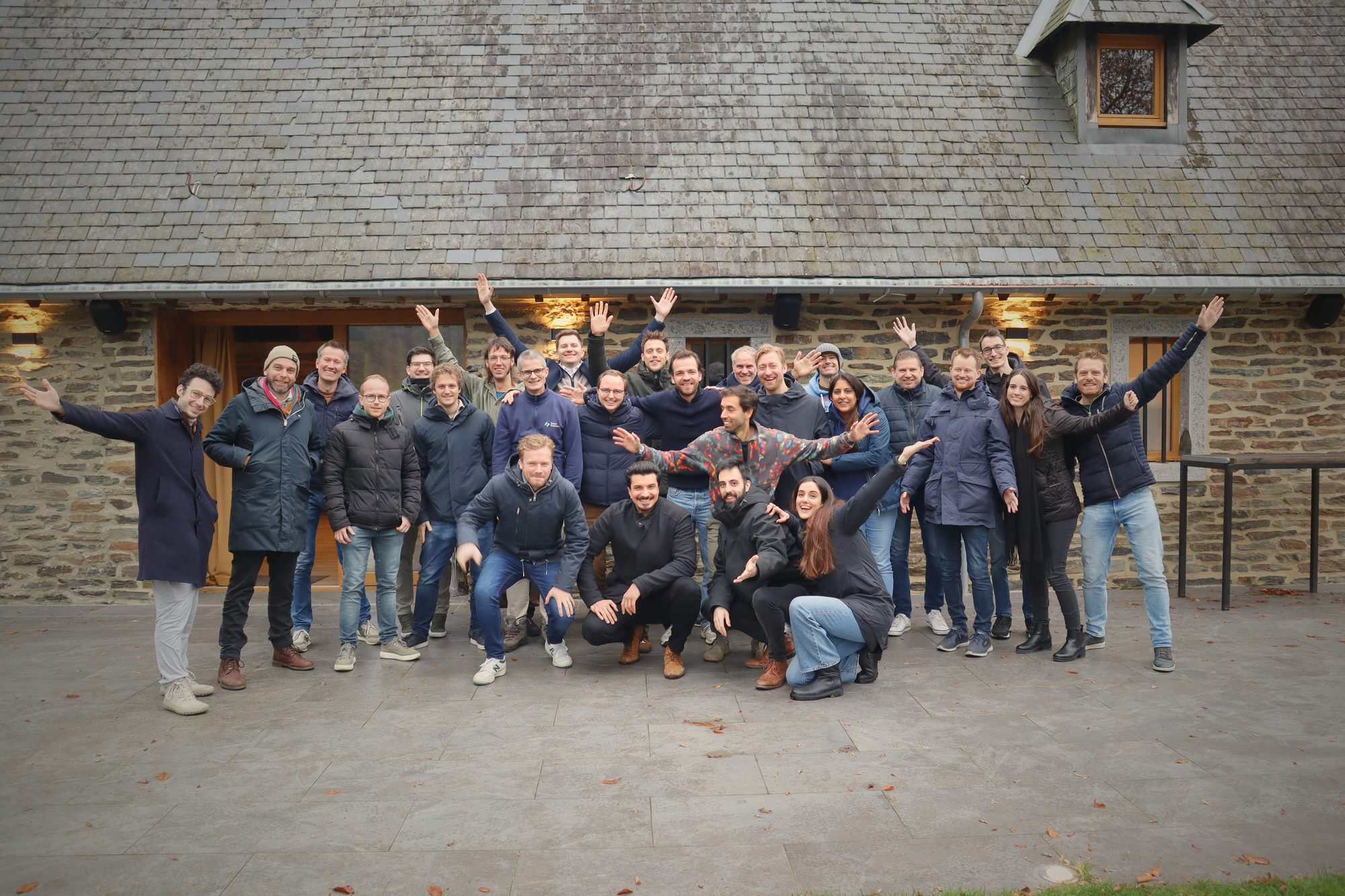 The Smart Robotics team, about thirty people, posing and waving in front of a stone building.